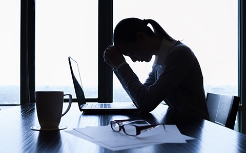 Silhouette of an office worker resting her head in her hands at a desk beside a laptop, a coffee mug and reading glasses