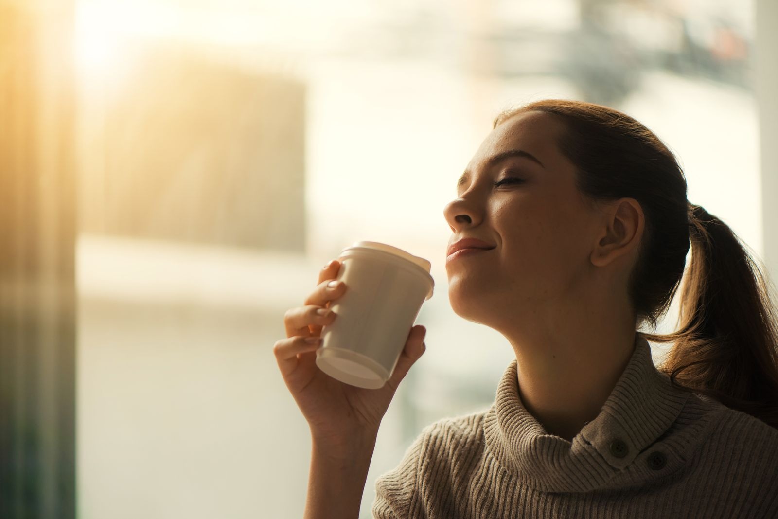 A woman in a soft jumper, eyes closed and smiling, holding a cup of tea in warm golden light